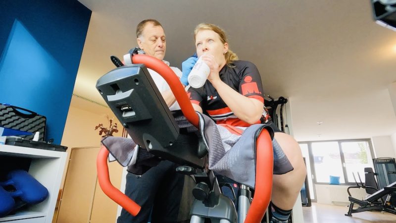 Athletin auf Fahrrad bei der Blutabnahme am Ohr während der Leistungsdiagnostik mit Trainer